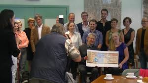 Peter Vanhoutte (voorzitter van Anna Timmerman vzw) overhandigt de cheque aan Nicole Kort (voorzitter van SWODB). Groepsfoto met de bestuursleden van Anna Timmerman vzw en de initiatiefnemers die het geld inzamelden.